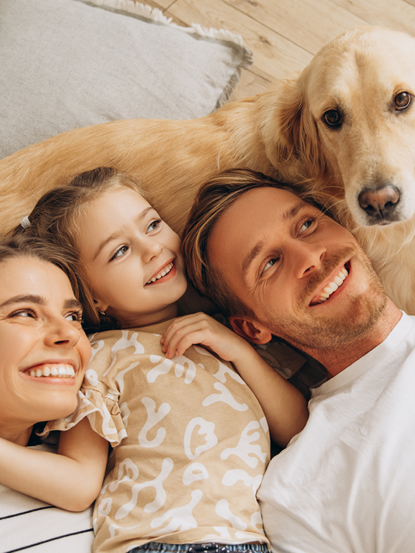 Eine junge Familie liegt auf dem Boden. Vater, Mutter und Tochter blicken den Hund an, welcher in die Kamera sieht.